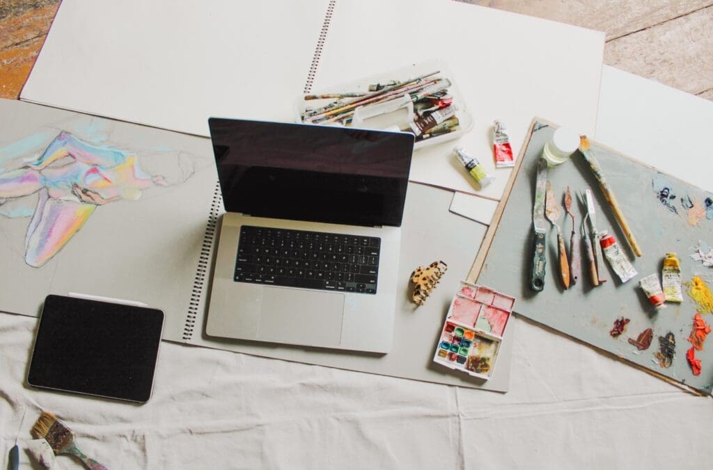 Flat lay of ipad, macbook, plus art products, tubes of paint, paint brushes and sketchbooks